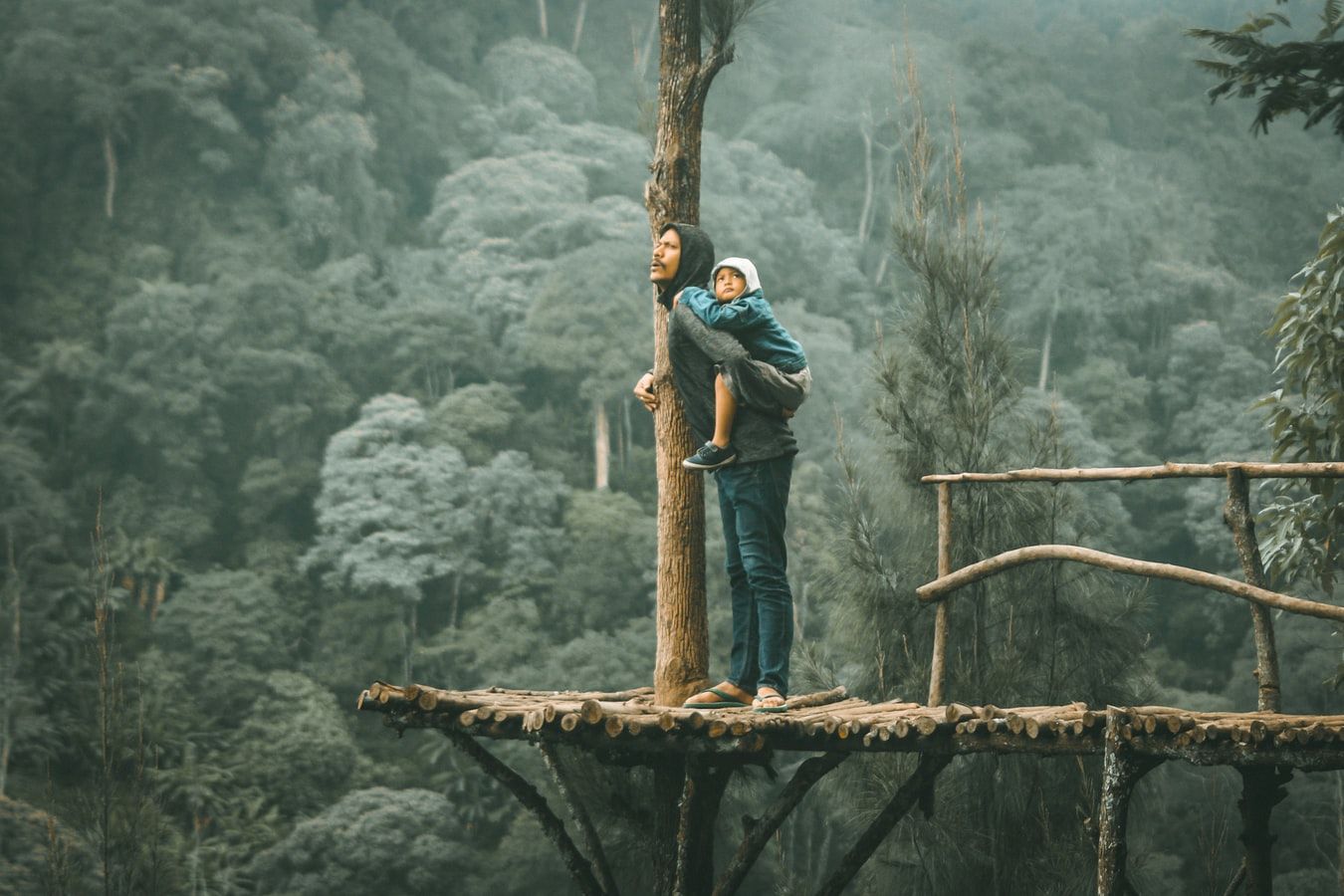 a woman is carrying a child on her shoulders while standing on a wooden bridge in the woods .