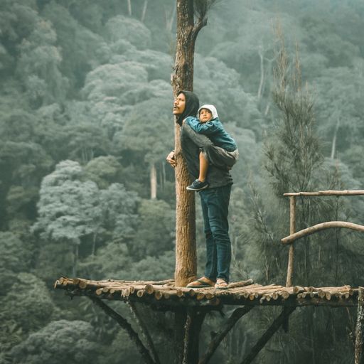 a woman is carrying a child on her shoulders while standing on a wooden bridge in the woods .