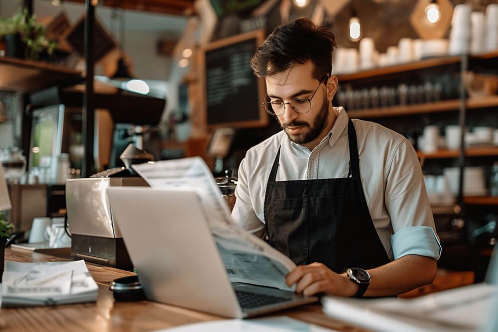 a man in an apron is using a laptop computer in a restaurant .