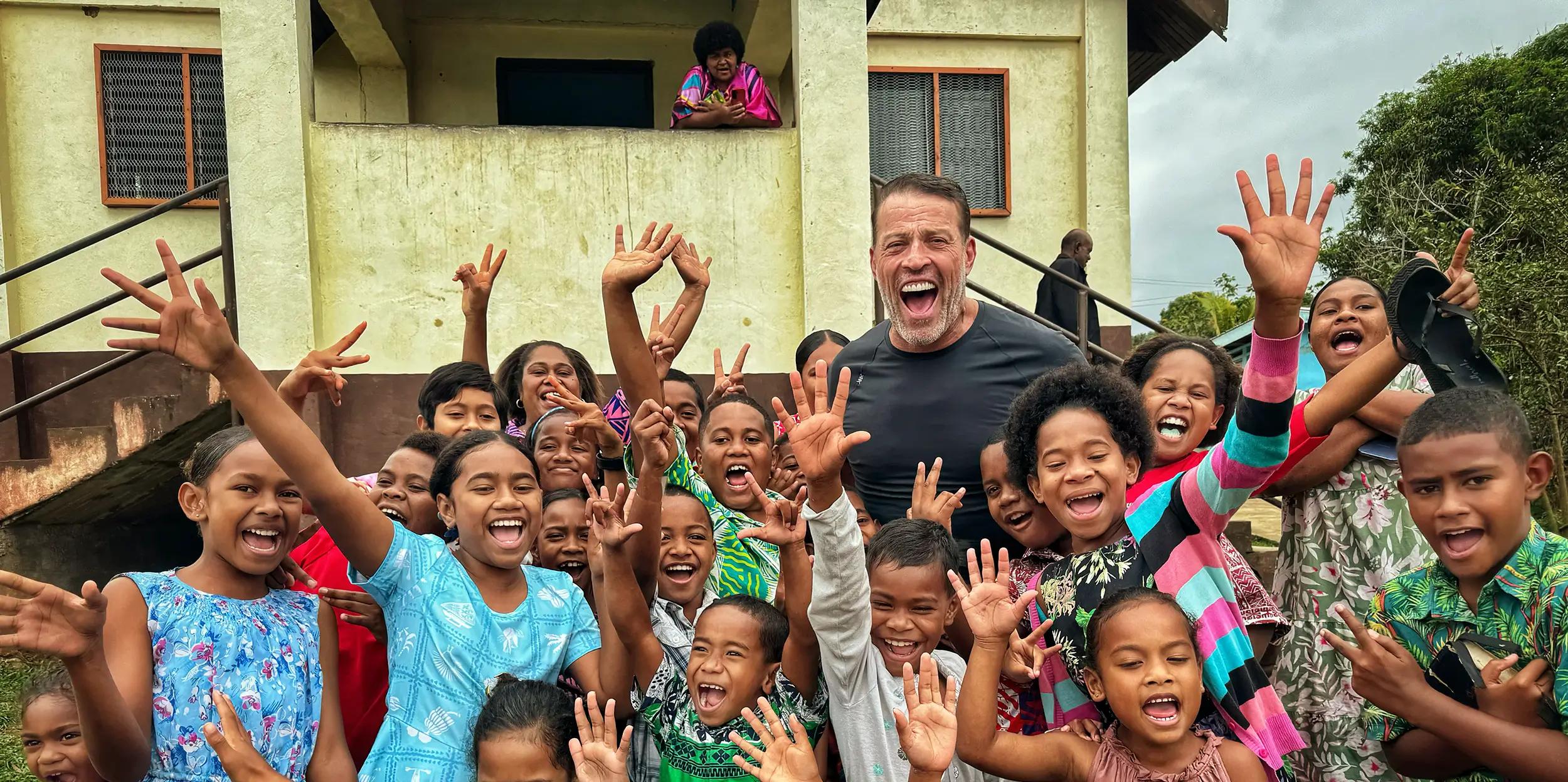 Tony Robbins and a large group of smiling children wave enthusiastically.