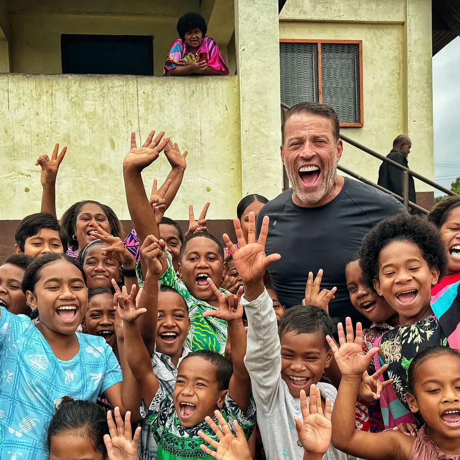 Tony Robbins and a large group of smiling children wave enthusiastically.