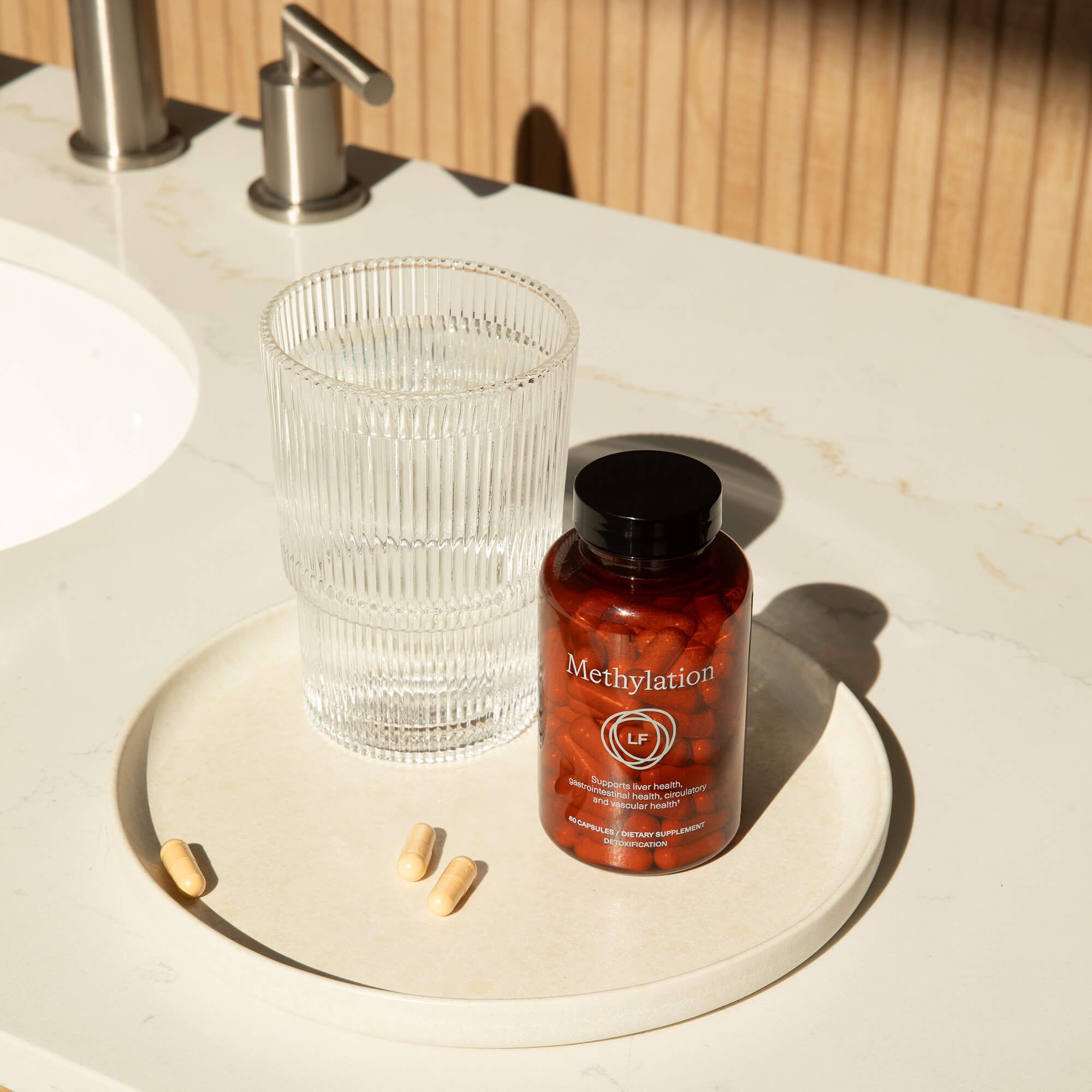 A bottle of "Methylation" supplements, a glass of water, and three capsules sit on a tray next to a sink.