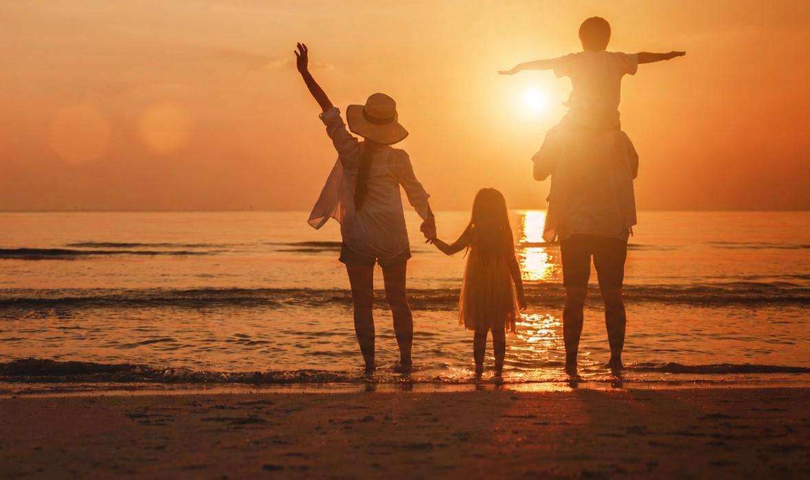 Silhouetted family of four on a beach at sunset, with a child on shoulders and another holding hands.