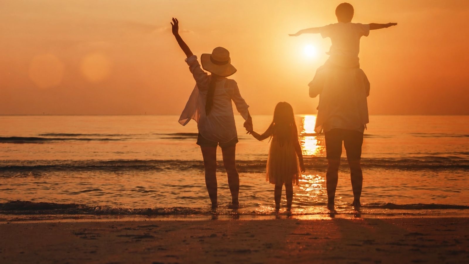 Silhouetted family of four on a beach at sunset, with a child on shoulders and another holding hands.