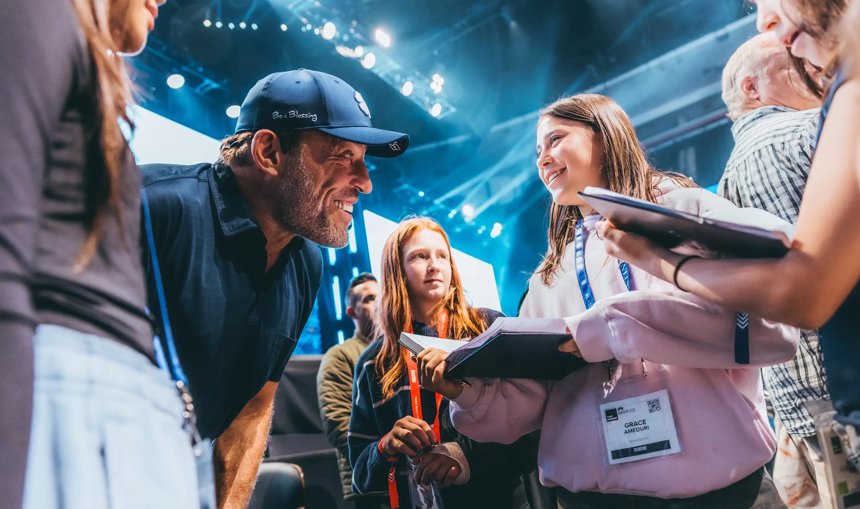 Tony Robbins, wearing a cap, smiles and leans toward a smiling young girl holding a book.