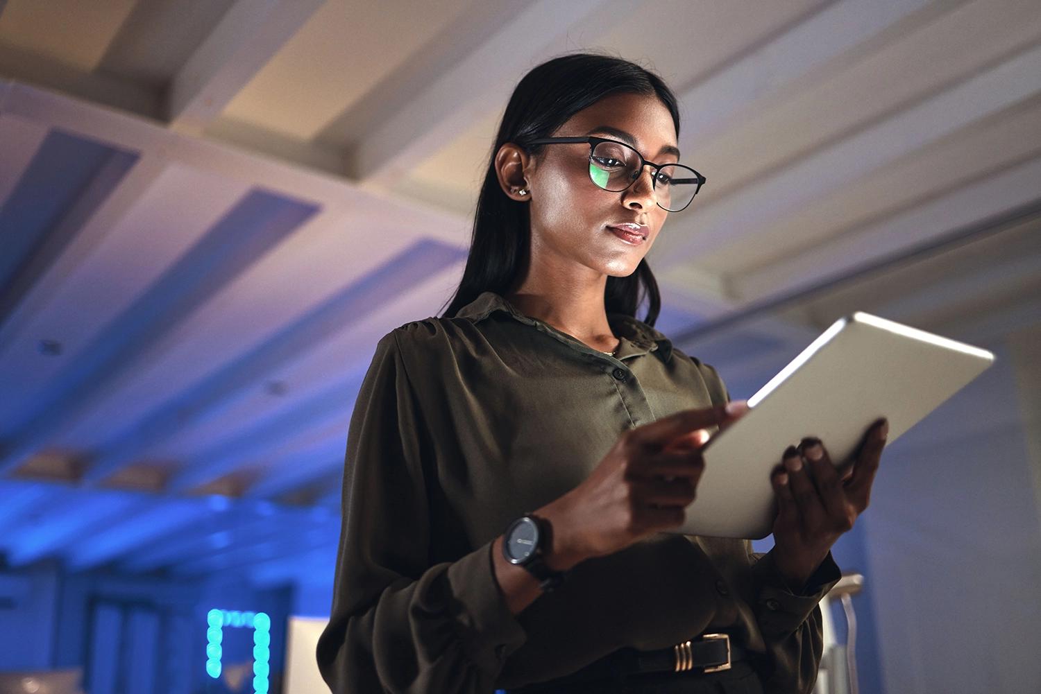 A woman in glasses, illuminated by her tablet screen, taps the screen.