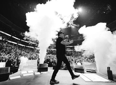 A man in black strides across a stage amid plumes of smoke, with a large audience visible in the background.