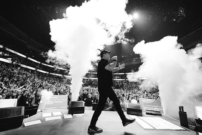 A man in black strides across a stage amid plumes of smoke, with a large audience visible in the background.
