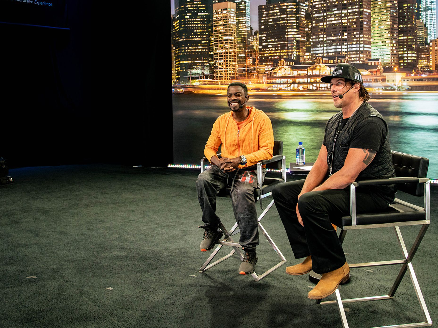 two men are sitting in director's chairs in front of a city skyline displayed on a screen