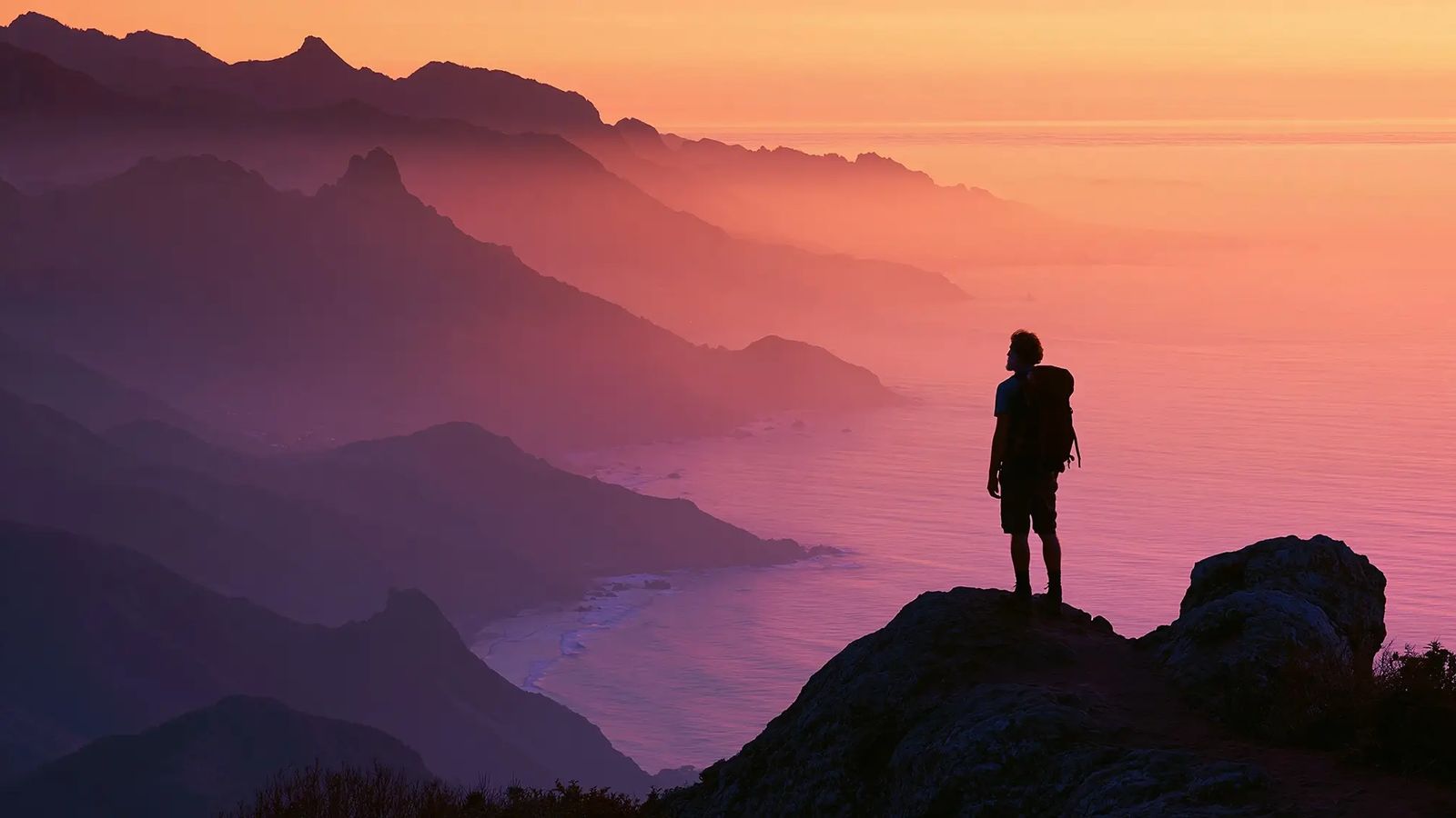 A hiker stands on a mountain peak overlooking a misty ocean and layered mountains at sunset.