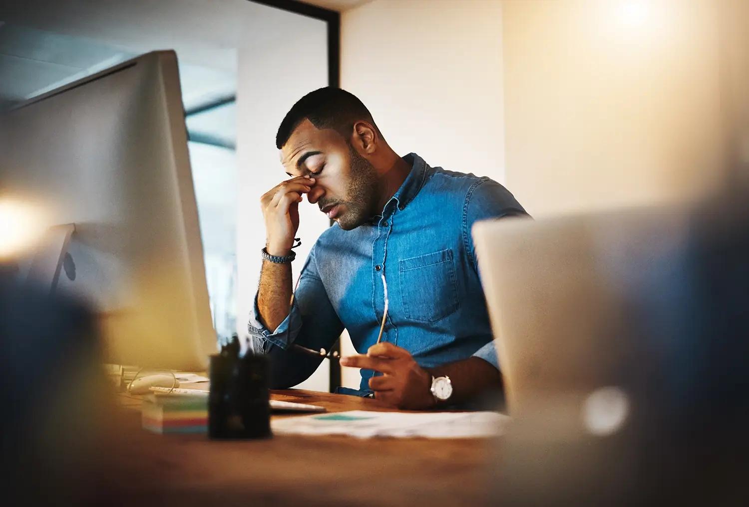 A man rubs his tired eyes while working at a computer desk.