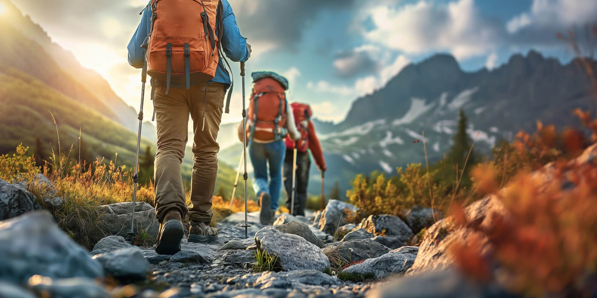 Three hikers with backpacks and trekking poles walk a rocky mountain trail under a warm sunset.