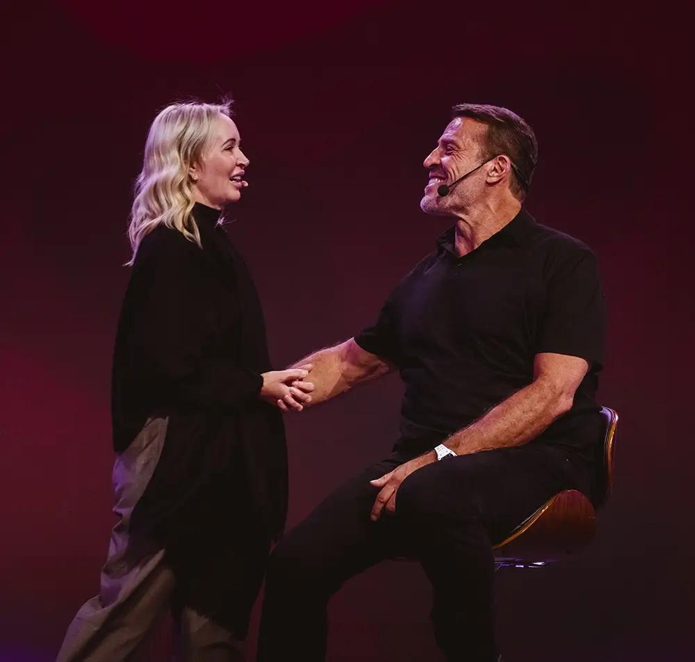 Tony Robbins, wearing a cap and lavalier mic, holds his hands over his chest with a heartfelt smile against a purple background.