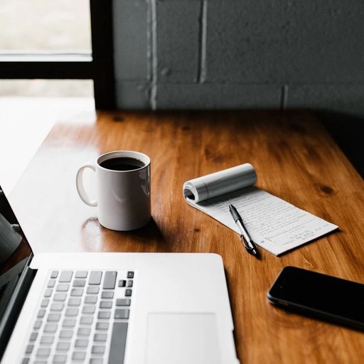 a wooden desk with a laptop , cup of coffee , notebook , pen and cell phone .