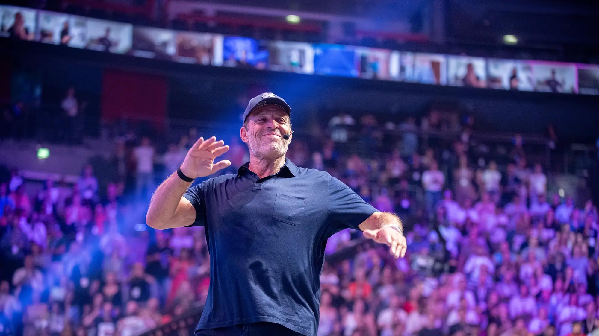 Tony Robbins smiling and gesturing to a large audience in an arena.