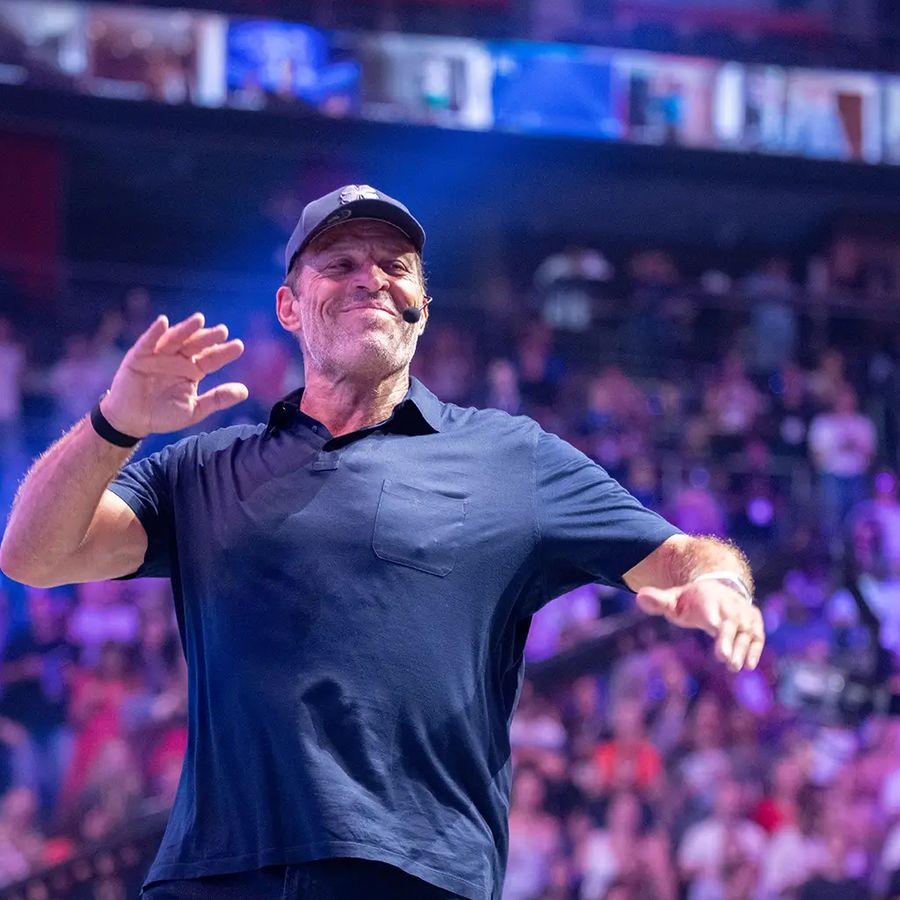 Tony Robbins smiling and gesturing to a large audience in an arena.