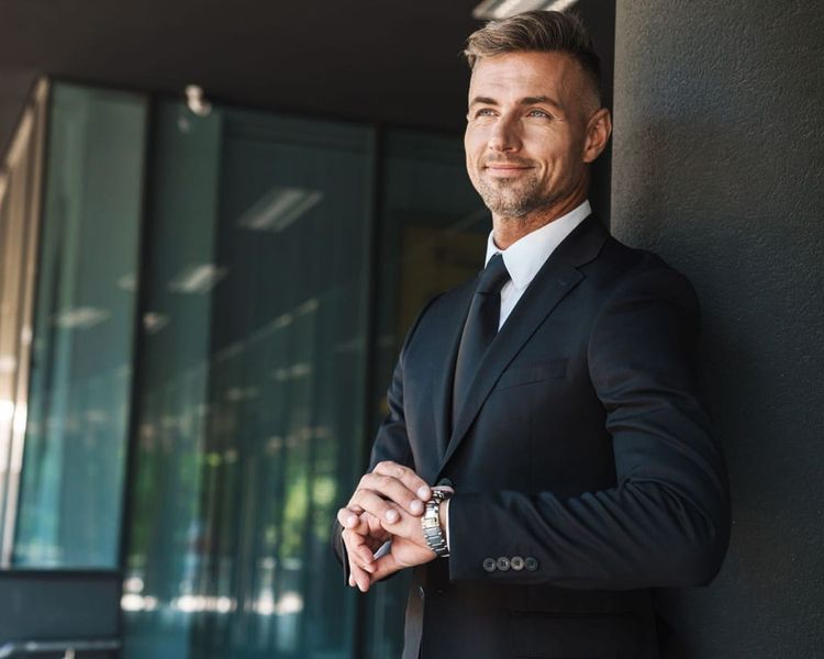 A smiling man in a black suit checks his watch, leaning against a dark pillar outdoors.