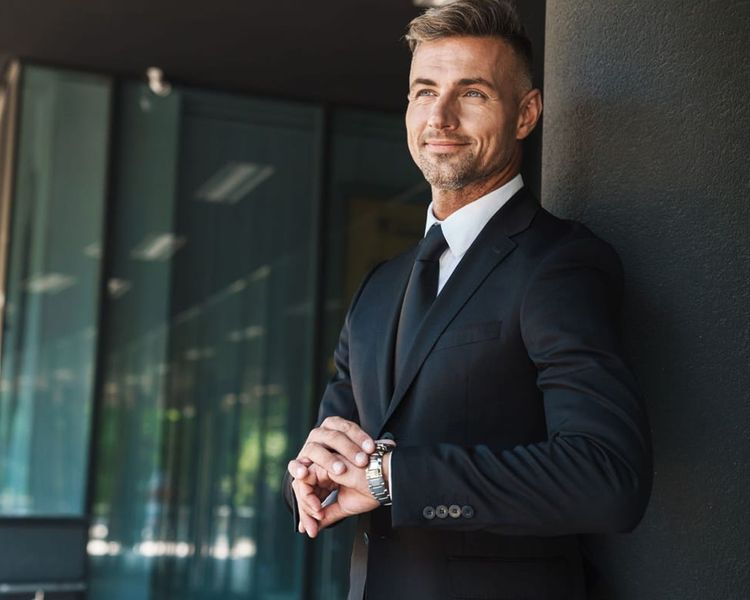 Smiling man in a suit checking his watch outside a modern building.