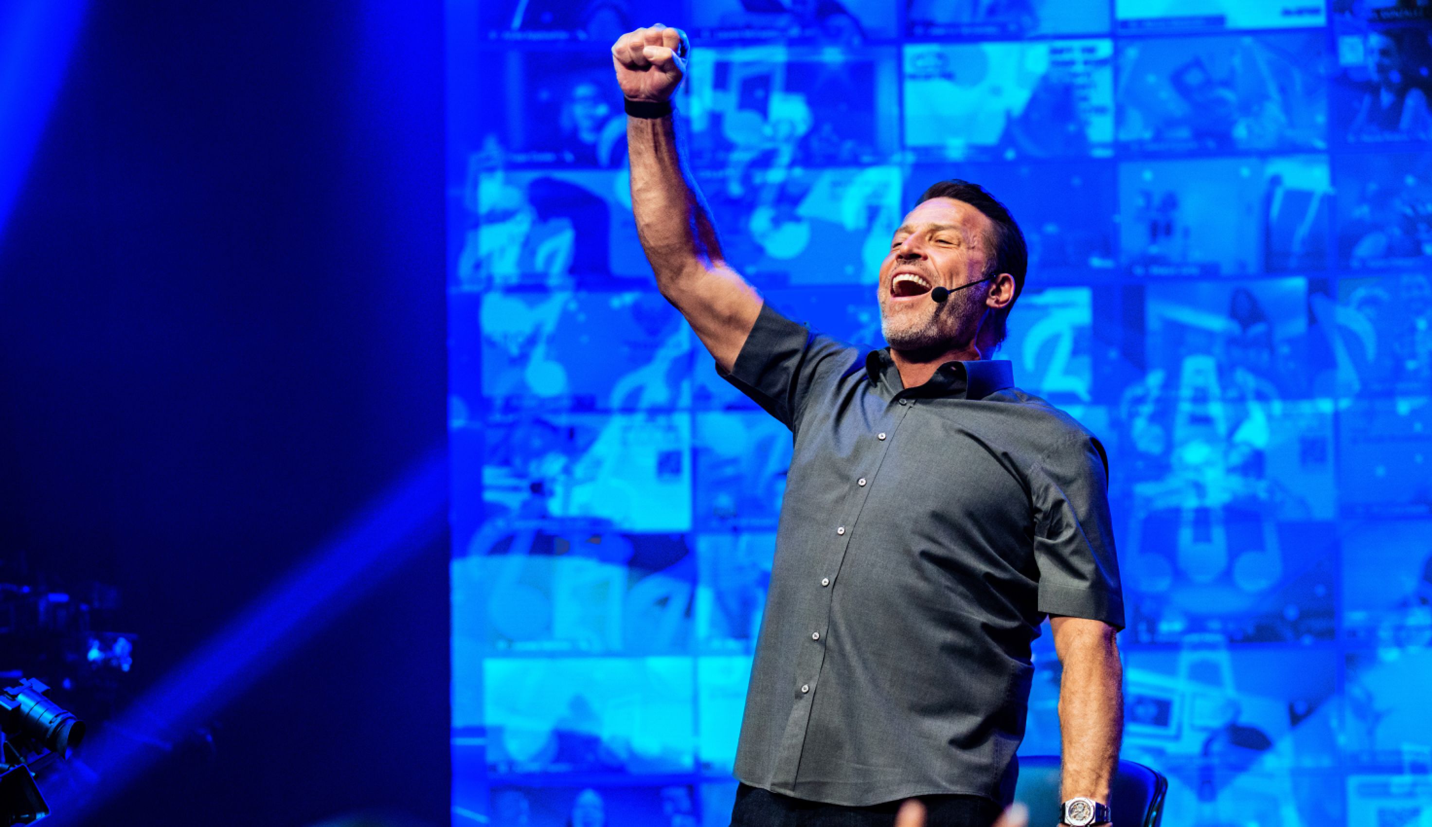 A man with a headset mic raises a triumphant fist and shouts on a blue-lit stage with screens in the background.