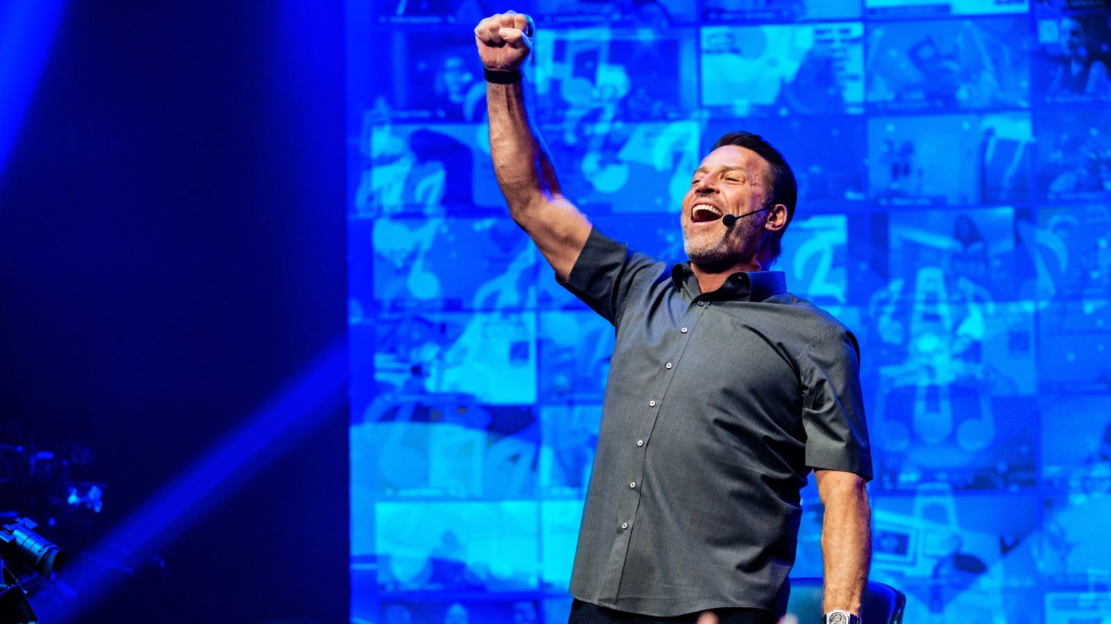 A man with a headset mic raises a triumphant fist and shouts on a blue-lit stage with screens in the background.