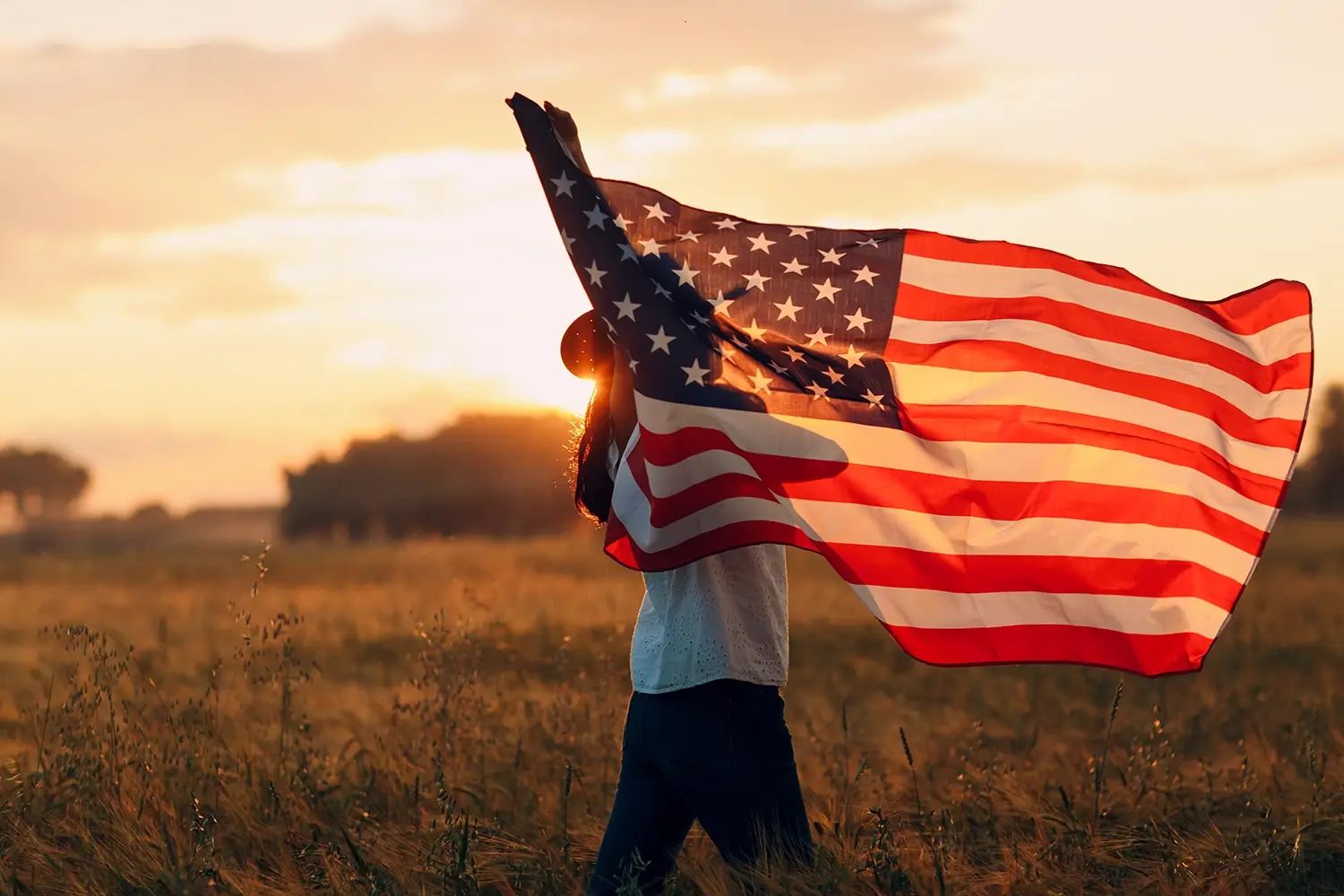 Person holding an American flag in a field at sunset.