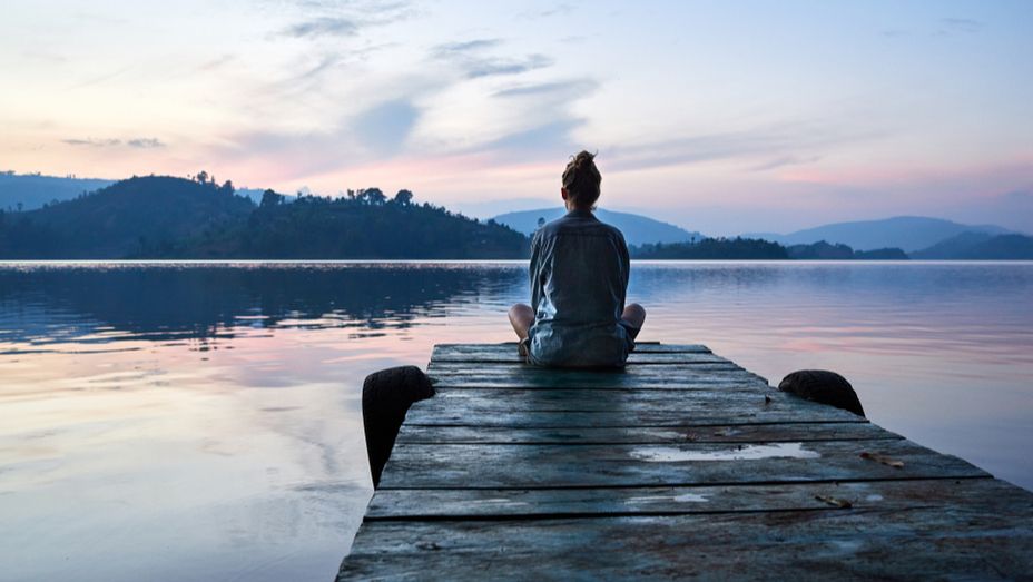 A person sits cross-legged on a wooden dock, facing a calm lake and mountains at sunset.