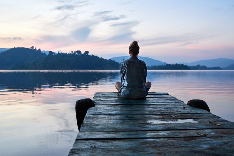 A person sits cross-legged on a wooden dock, facing a calm lake and mountains at sunset.