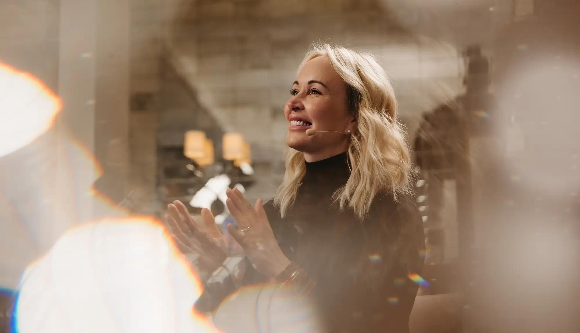A smiling blonde woman with a microphone claps, surrounded by warm lens flares.