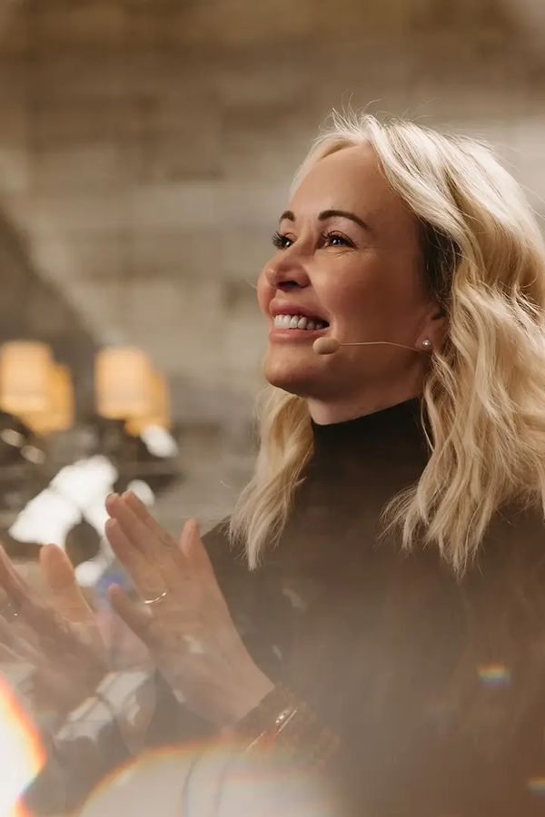 A smiling blonde woman with a microphone claps, surrounded by warm lens flares.