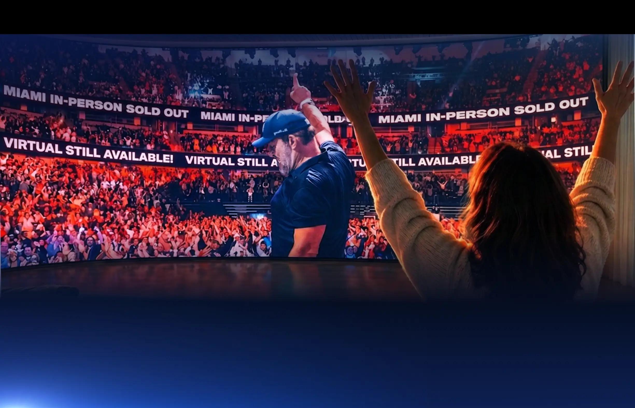 Three people from behind watch a large screen showing a man on stage waving to a cheering crowd, with the viewers also raising their hands.