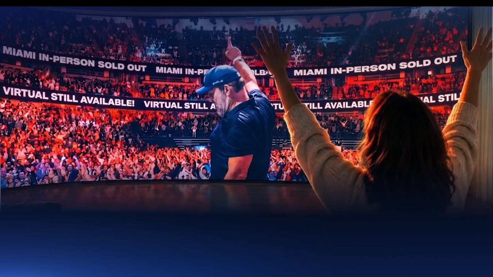 Three people from behind watch a large screen showing a man on stage waving to a cheering crowd, with the viewers also raising their hands.