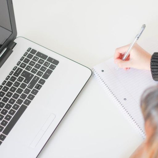 a person is writing in a notebook in front of a laptop computer .
