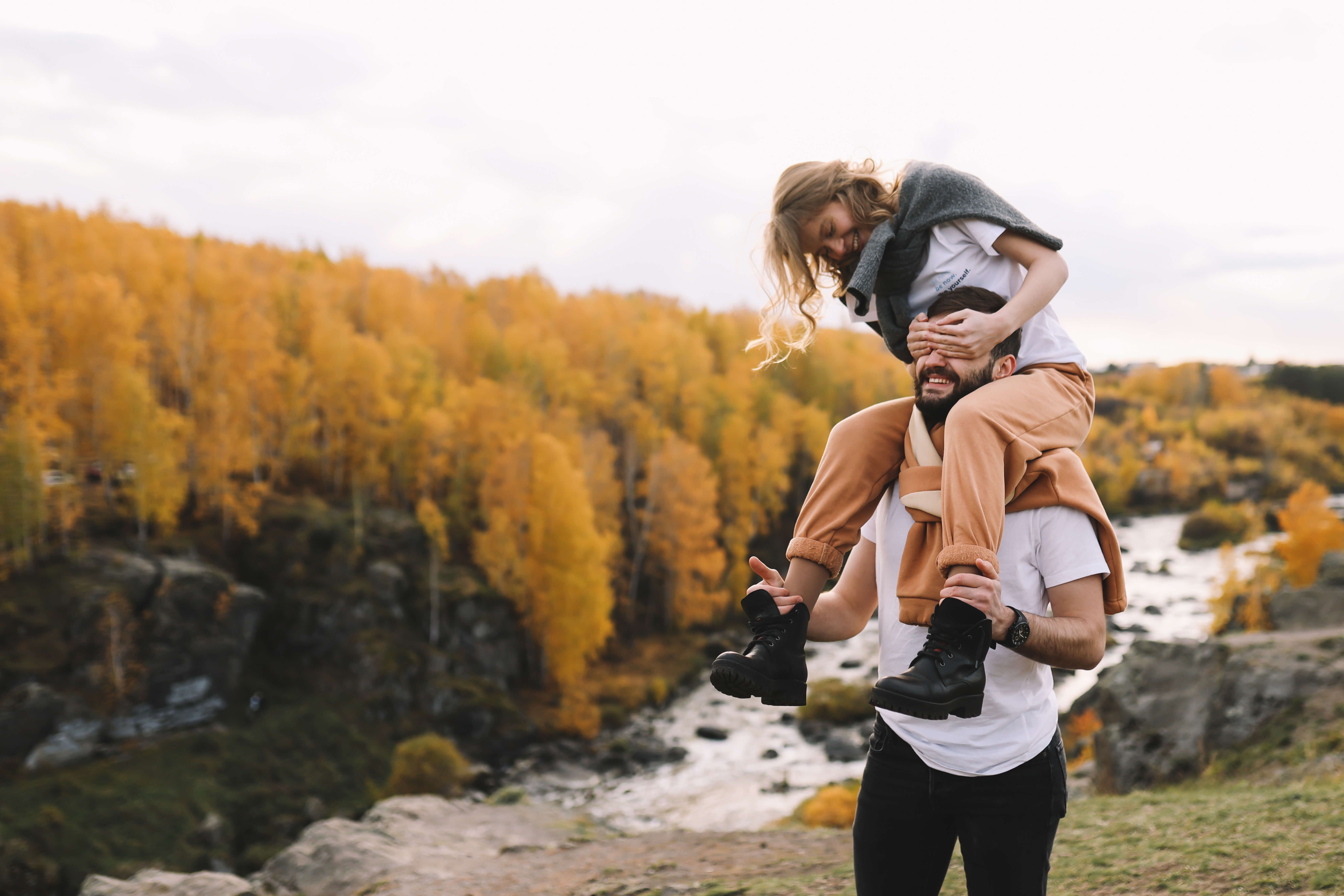 A woman sits on a man's shoulders, covering his eyes, in a vibrant autumn forest landscape with a river below.