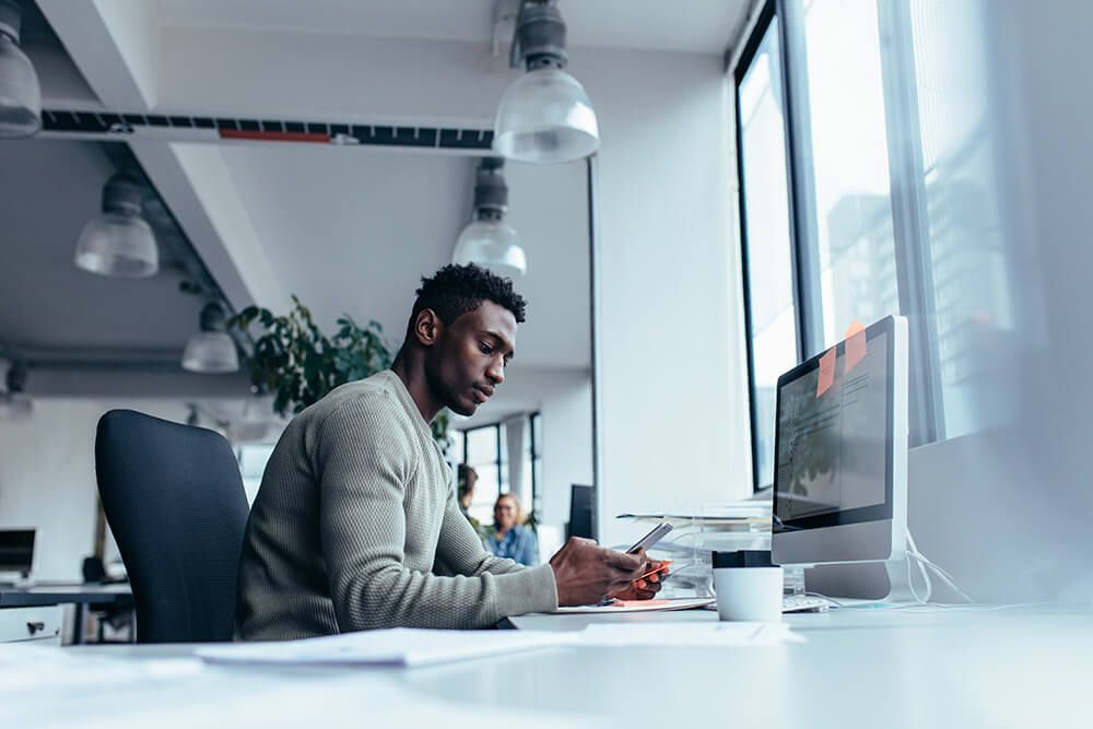 a man is sitting at a desk in front of a computer using a cell phone .