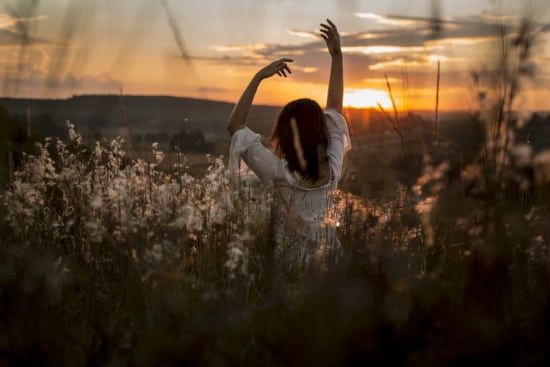 a woman is standing in a field of tall grass with her arms outstretched at sunset .