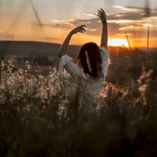 a woman is standing in a field of tall grass with her arms outstretched at sunset .