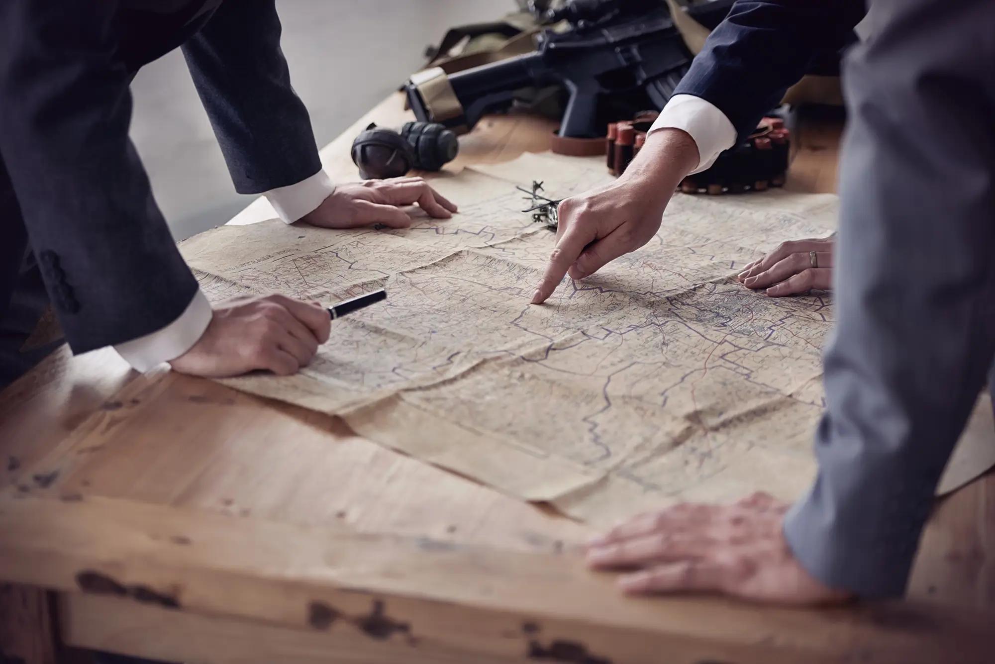 Hands of people in suits pointing at a map with military equipment nearby.