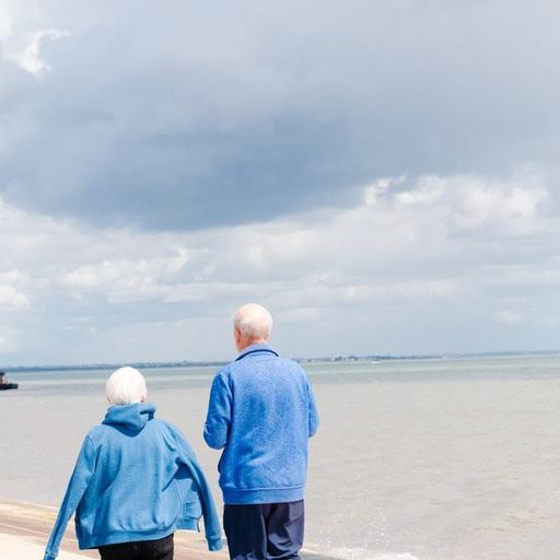 an elderly couple is walking along the beach holding hands .