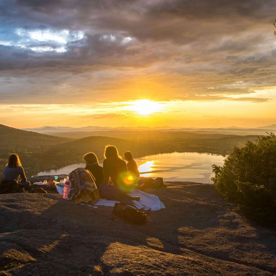 a group of people are sitting on top of a rock watching the sunset over a lake .