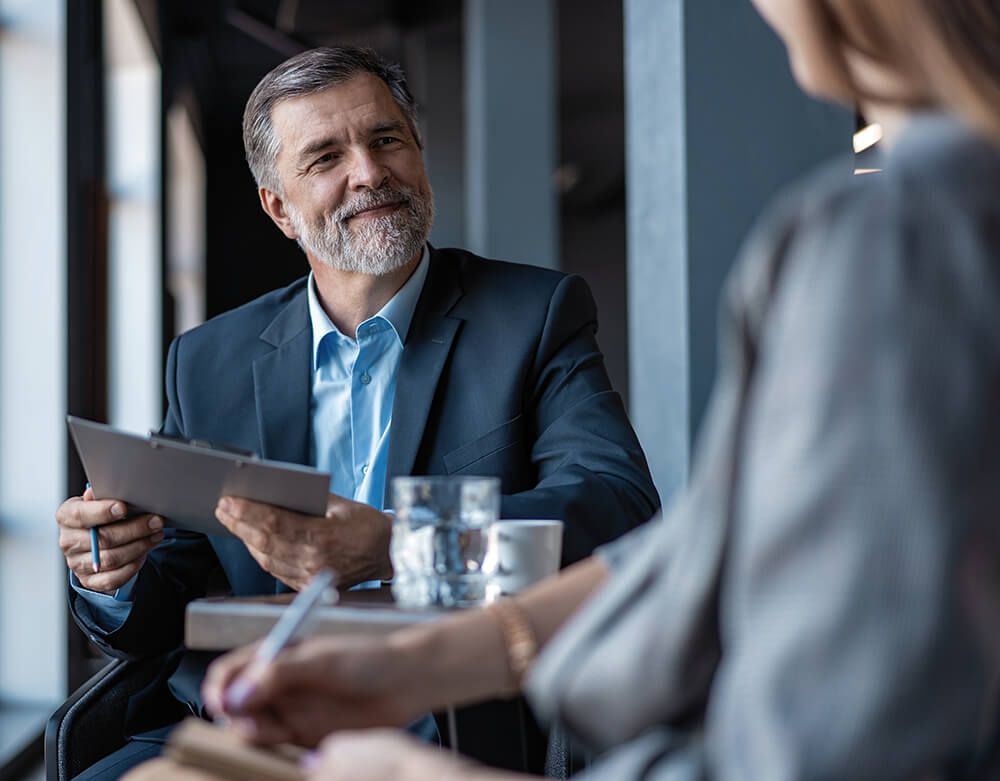 a man in a suit is sitting at a table with a woman and holding a clipboard .