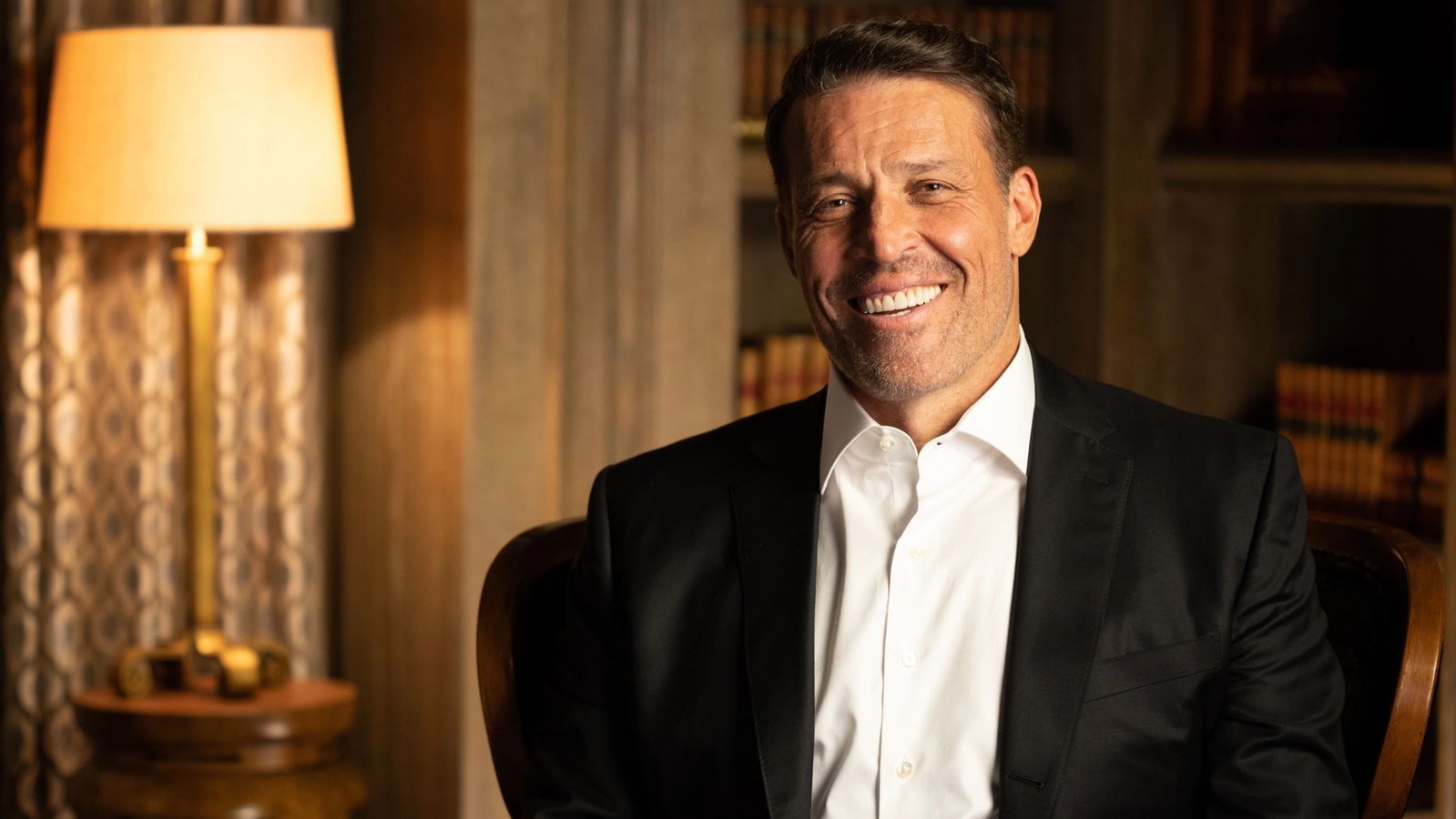 a man in a suit and white shirt is sitting in a chair and smiling .