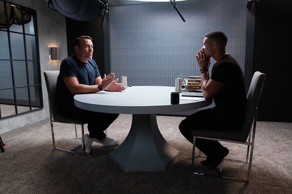 Two men sit at a round table, one gesturing as he speaks while the other listens intently.