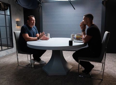 Two men sit at a round table, one gesturing as he speaks while the other listens intently.
