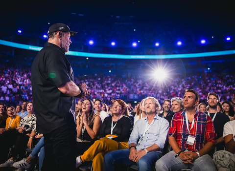 Speaker on stage addressing a large, attentive audience in an arena.