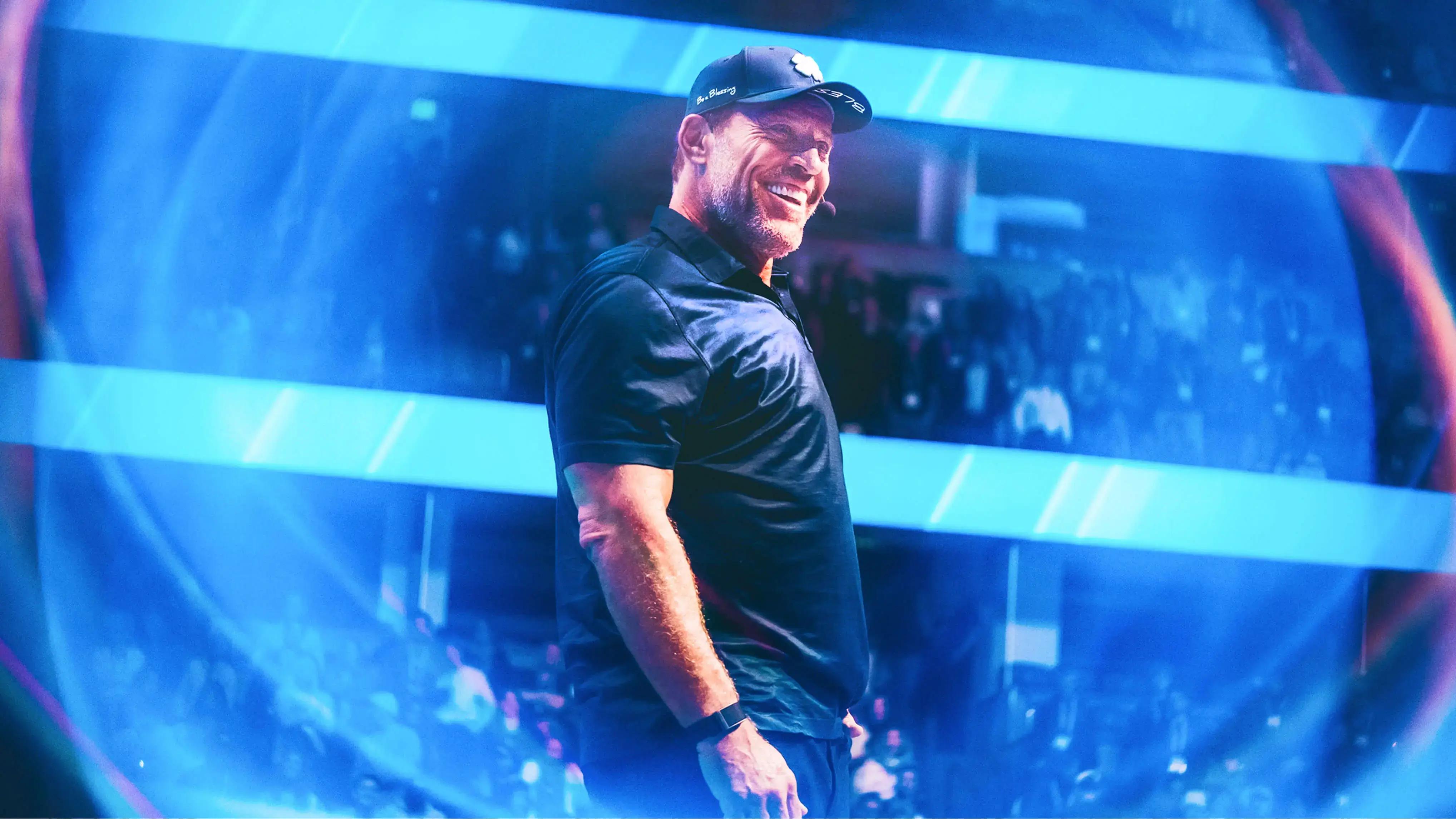 A smiling man in a black cap and shirt on a stage with a blue, futuristic glow.
