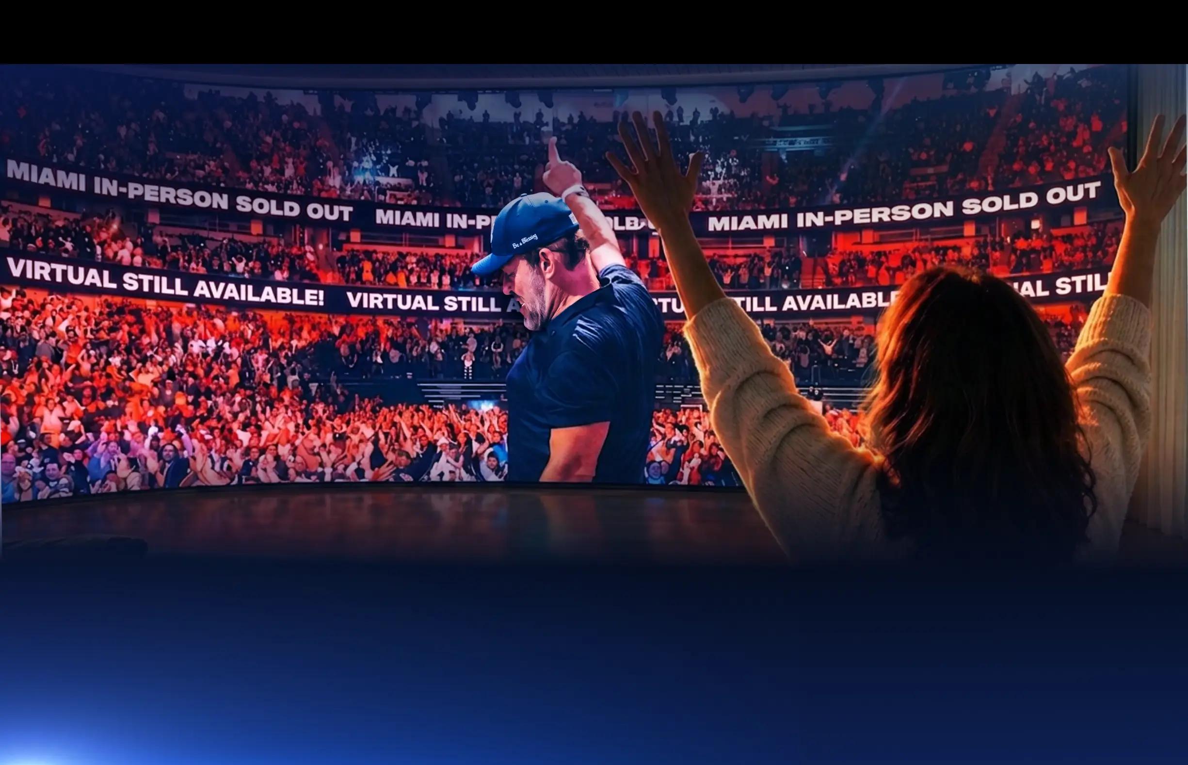 Three people from behind watch a large screen showing a man on stage waving to a cheering crowd, with the viewers also raising their hands.