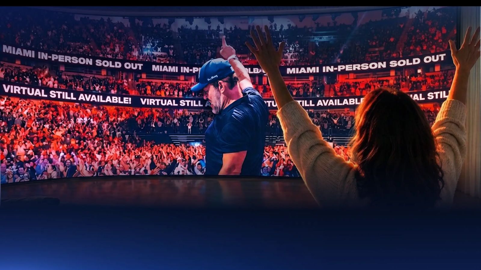 Three people from behind watch a large screen showing a man on stage waving to a cheering crowd, with the viewers also raising their hands.