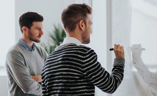 a man is writing on a whiteboard while another man looks on .