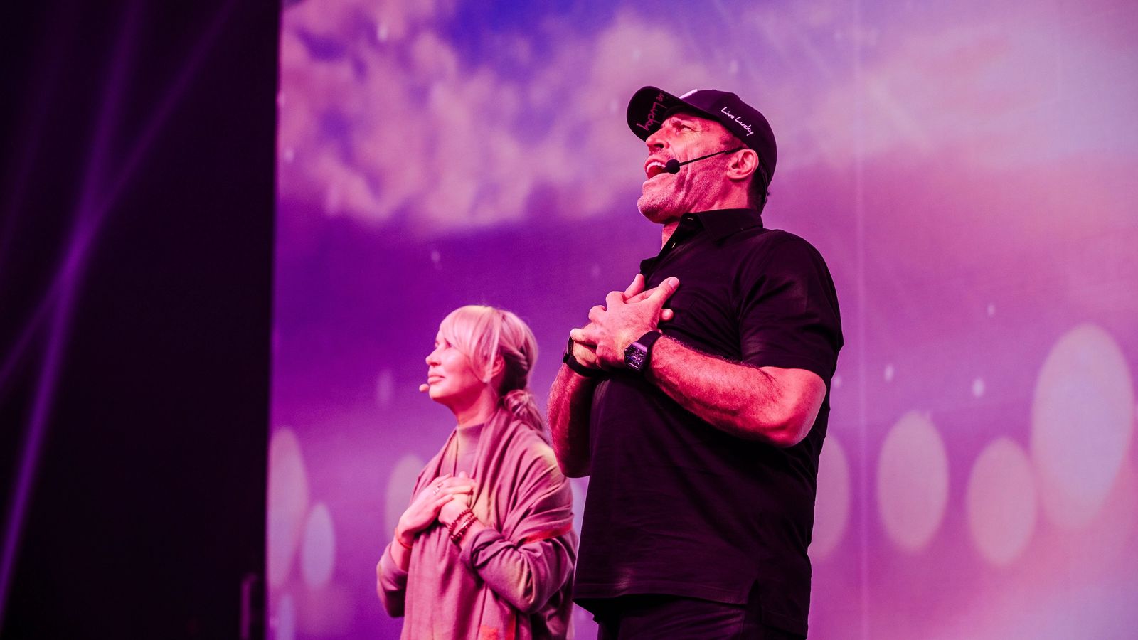 Tony Robbins and a woman on stage, hands over hearts, looking up under purple lighting.
