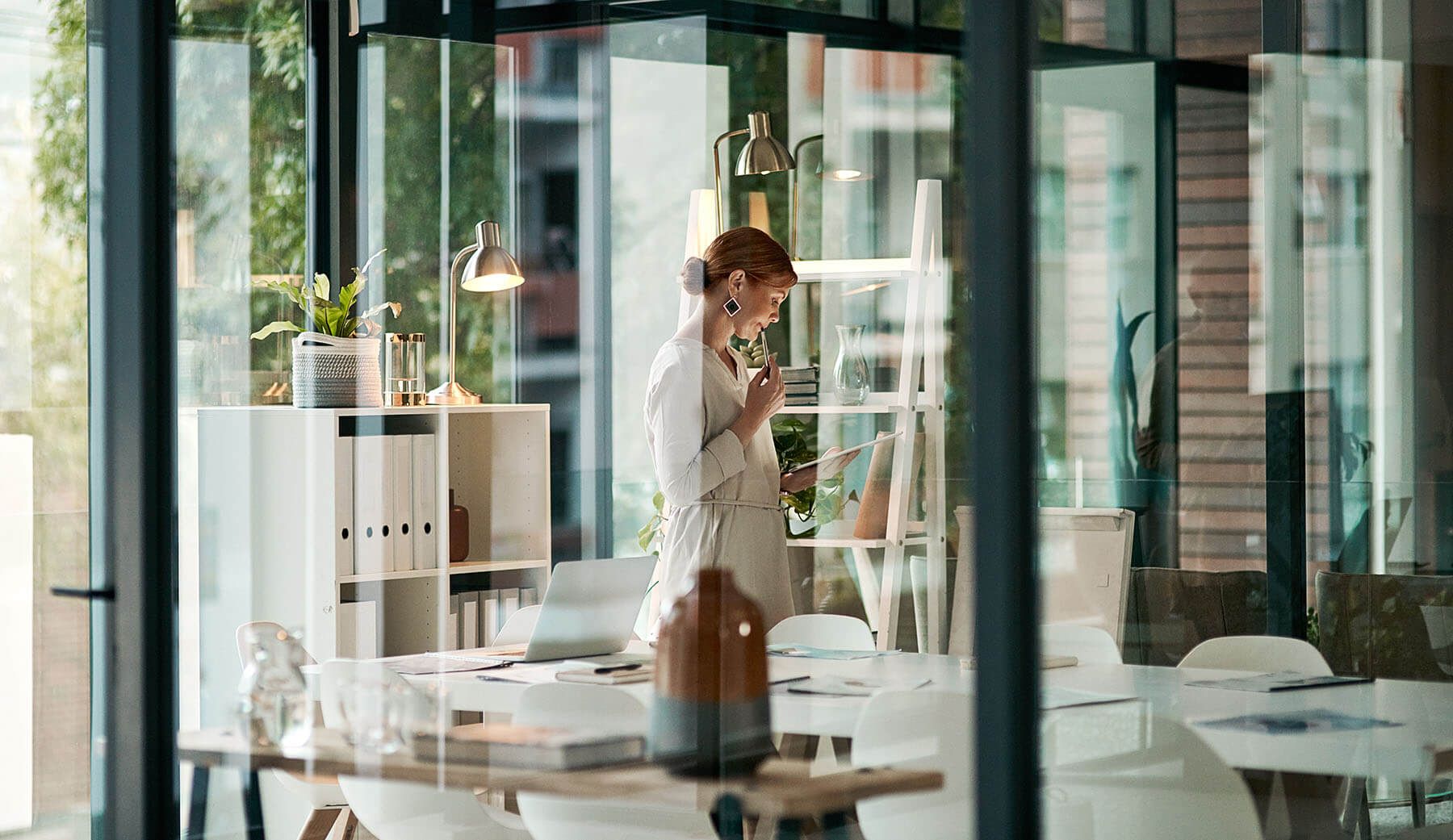 Thoughtful woman reviewing documents in a modern office, viewed through glass.